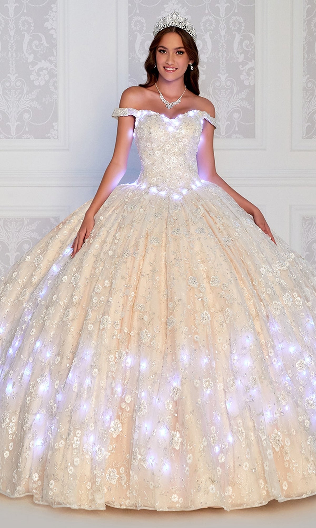 A young woman in the Princesa Glow-in-the-Dark Quince Dress PR12266, accessorized with a tiara and necklace, stands before a white decorative wall as her dress glows with sparkling lights.